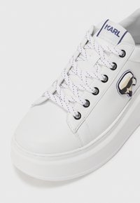 White leather sneaker with textured surface, purple-accented laces, and a cartoon logo. Features a thick platform sole for added height.