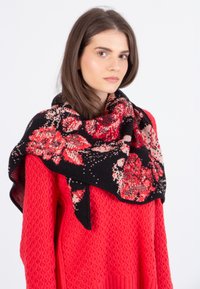 Gehaakte bloemen sjaal in zwart met rode en beige patronen, losjes over een rode textuurtrui gedrapeerd. Zacht materiaal met een gezellige afwerking.