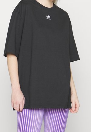 Osoba ubrana w prostą, czarną, oversize koszulkę Adidas z logo na piersi i fioletowo-białe paskowane spodnie, stojąca na jednolitym tle.
