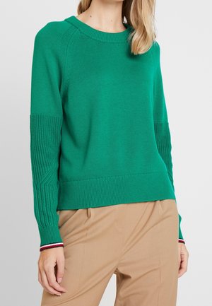 Femme portant un pull en maille vert vif avec des manches texturées et un pantalon beige sur un fond clair uni.