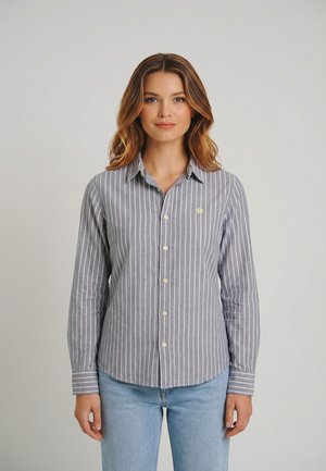 Joven mujer de pie, vestida con una camisa gris de botones con rayas verticales blancas y jeans azul claro, frente a un fondo liso.