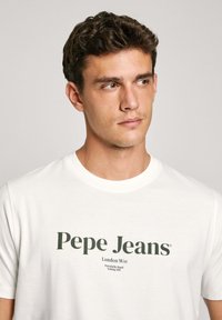 Camiseta de algodón blanca con texto verde que dice "Pepe Jeans London W11" y un subtítulo más pequeño. Mangas cortas y cuello redondo.