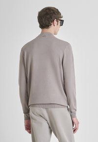 Uomo che indossa un maglione taupe a maniche lunghe e pantaloni grigi chiari, in piedi e rivolto di schiena con occhiali da sole neri, su uno sfondo bianco semplice.