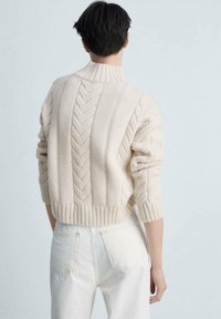 Cremfarbener Kabelstrickpullover mit hohem Kragen, strukturierten Mustern und geripptem Saum; kombiniert mit hellen Jeans.
