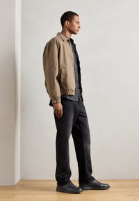 Giacca bomber marrone chiaro, maglietta a righe scure, jeans neri e scarpe grigie, in piedi su un pavimento di legno con uno sfondo semplice.