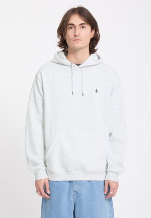 Jeune homme aux cheveux bruns portant un sweat à capuche gris clair et un jean bleu clair, debout devant un fond blanc uni.