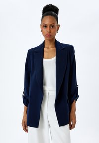 Blazer blu navy in tessuto leggero con colletto a punta, apertura frontale e maniche con risvolto. Indossato sopra una maglietta bianca e pantaloni chiari.