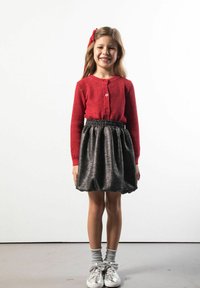 Młoda dziewczynka uśmiecha się, ubrana w czerwony sweter, metaliczną czarną spódnicę, szare skarpetki, srebrne buty z kokardkami oraz czerwoną opaskę na głowę na jednolitym tle.