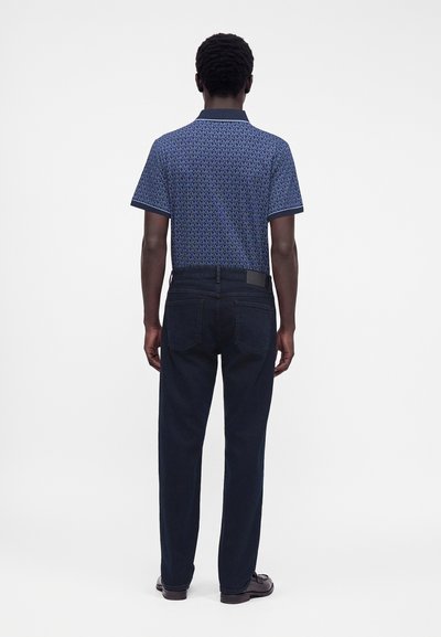 Homme vu de dos portant un polo bleu à manches courtes à motifs, un jean foncé et des chaussures foncées sur un fond uni.