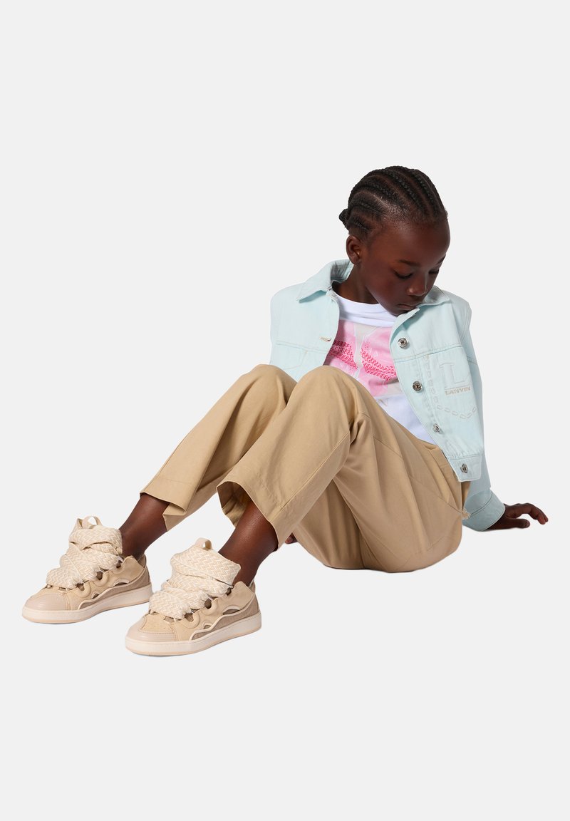 Barn med flettet hår, siddende iført en lyseblå jakke, hvid og pink trøje, beige bukser og beige højtop-sneakers med tykke snørebånd.