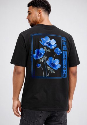 Mann trägt schwarzes T-Shirt mit blauen Anemonenblumen und der Aufschrift „LUMINOUS“ auf dem Rücken vor einfarbigem Hintergrund.