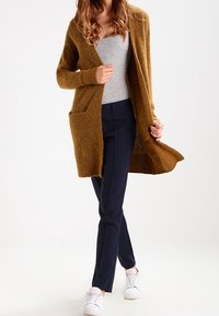 Cardigan largo color mostaza hecho de un tejido texturizado. Lleva puesto sobre una camiseta gris y se combina con pantalones azul marino y zapatillas blancas. Tiene bolsillos delanteros.