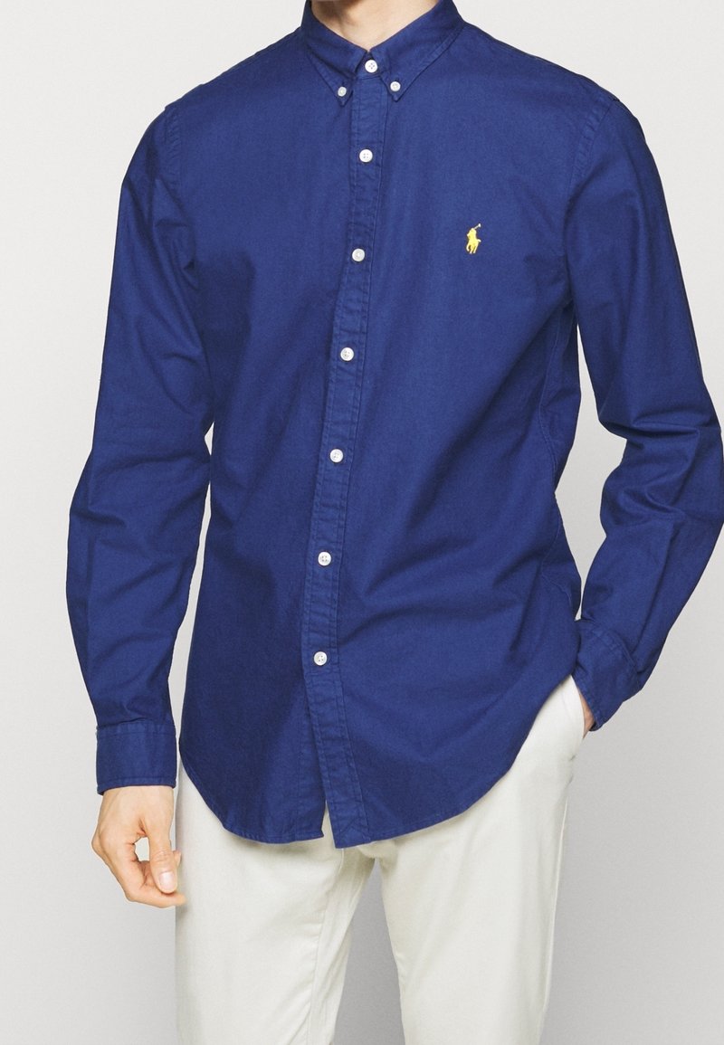 Mies pukeutuneena napitettuun tummansiniseen pitkähihainen paitaan, jossa on keltainen kirjailtu logo, ja vaaleat housut, käsi taskussa.