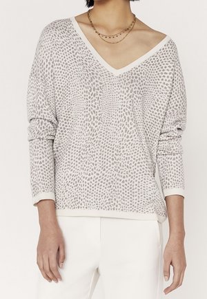 Femme portant un pull à col en V à motifs gris clair à manches longues et un pantalon blanc, accessoirisée de colliers dorés superposés.