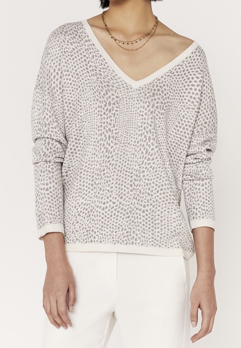 Femme portant un pull à col en V à motifs gris clair à manches longues et un pantalon blanc, accessoirisée de colliers dorés superposés.