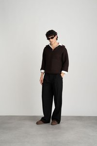 Suéter marrón de punto acanalado con una camisa blanca de cuello, combinado con pantalones negros de pierna ancha y zapatos marrones sin cordones. Las gafas de sol completan el look.