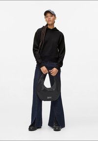 Jovem mulher vestindo um moletom preto, jeans flare escuros com fendas na frente, botas pretas, boné cinza, segurando uma bolsa preta com logotipo em frente a um fundo branco.