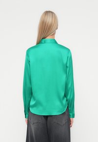 Camisola longa de mangas em satin verde com gola. Textura lisa, bainha ligeiramente curva e punhos com botão. Usada com calças cinzentas escuras.