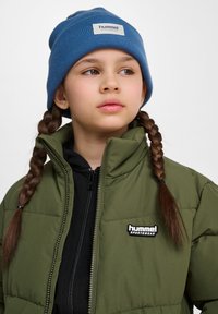 Grön puffjacka med svart dragkedja, med en applikation som säger "hummel SPORTSWEAR," i kombination med en blå stickad mössa.
