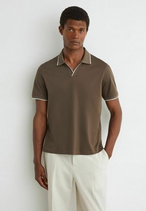Mann trägt ein braunes kurzärmeliges Poloshirt mit weißem Besatz und hellbeiger Hose, steht vor einer weißen Wand.