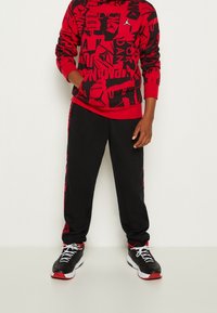 Punainen ja musta kuviollinen huppari kengurutaskulla, yhdistettynä mustiin sweattihousuihin, joissa on punaiset sivuraidat. Mustat lenkkarit punaisilla yksityiskohdilla.