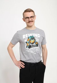 Šedé bavlněné tričko s grafickým potiskem, na kterém je karikaturní robot, text a abstraktní vzory; nošeno s černými kalhotami a opaskem.