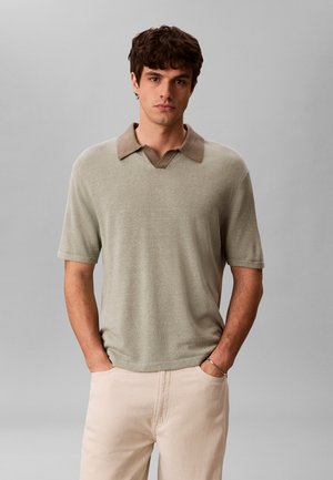 Muž s tmavými vlasmi nosí béžové krátky rukáv pletený sveter s hnedým golierom a svetlými nohavicami, stojí s rukami v vreckách.