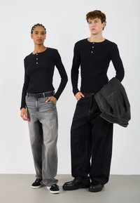 Dva modeli nosita črne dolge srajce s kratkimi rokavi. En ima na sebi lahke, svetleče sivo kavbojke in črn pas; drug pa nosi temno široke kavbojke in jakno.