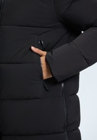 Zwarte pufferjack met ritssluitingen aan de zijkanten en op de borst, een gevoerde textuur en een aansluitend design met ribbelmanchetten.