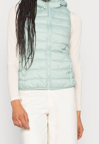 Light blue puffer vest with a zip front and hood. Features quilted design and a smooth, shiny texture. Worn over a fitted long-sleeve top.