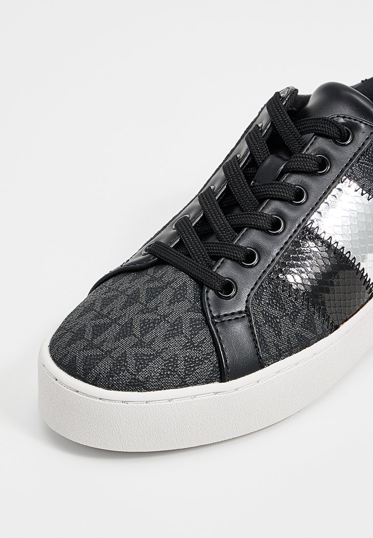 Zapatillas negras con una parte superior de tela texturizada, acentos negros brillantes, un patrón geométrico plateado en el lado y una suela de goma blanca.