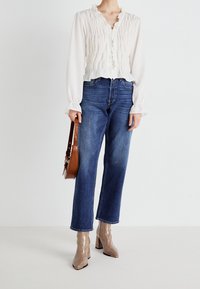 Bílá blůza s volánky a dlouhými rukávy, spárovaná s modrými zkrácenými džíny. Lesklé béžové kotníkové boty a hnědá kabelka přes rameno jsou součástí outfitu.