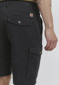 Zwarte cargo shorts met een zijzak en een achterzak met een knoop, voorzien van een beige label. Gemaakt van duurzame, stevige stof.