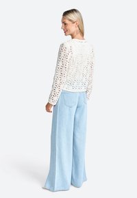 Maglione bianco all'uncinetto con motivo floreale, abbinato a jeans a gamba larga di un blu chiaro. Design testurizzato e vestibilità ampia. Nessun altro dettaglio visibile.