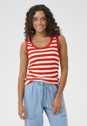 Uśmiechnięta kobieta o kręconych włosach nosi czerwono-biały, paskowany bezrękawnik oraz jasnoniebieskie spodnie ze sznurkiem na tle jednolitego tła.