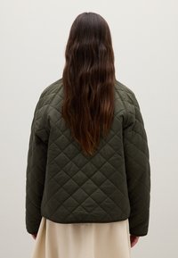 Person mit langen braunen Haaren, die eine dunkelgrüne gesteppte Jacke und einen beigen Rock trägt, steht vor einer schlichten hellgrauen Wand.