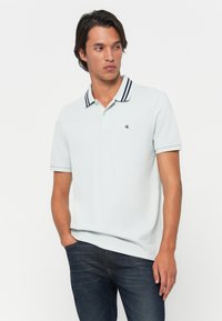 Mann iført lys blå polo med mørk stripet krage og blå jeans, stående med den ene hånden i lommen, ser sidelengs.