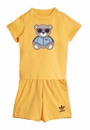 Żółty zestaw z koszulką z krótkim rękawem oraz szortami, na koszulce nadruk niedźwiedzia w okularach przeciwsłonecznych i niebieskiej kurtce, logo Adidas na szortach.