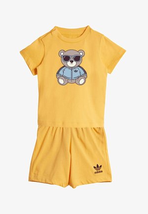 Żółty zestaw z koszulką z krótkim rękawem oraz szortami, na koszulce nadruk niedźwiedzia w okularach przeciwsłonecznych i niebieskiej kurtce, logo Adidas na szortach.