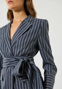 Harmaa pinstripe-takkia säädetyllä istuvuudella, syvällä V-kaula-aukolla ja sidottavalla vyöllä. Kankaan rakenne on teksturoitu ja siinä on sinisiä raitoja sekä jämäkkä kaulus.