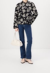 Czarna pikowana kurtka w kwiaty z okrągłym dekoltem, dwie kieszenie, zestawiona z rozkloszowanymi niebieskimi dżinsami i kremową torebką. Beżowe buty z paskami jako akcent.