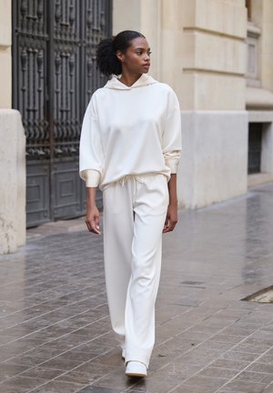Felpa bianca con cappuccio, dal taglio rilassato e coulisse, abbinata a pantaloni larghi coordinati. Indossata con scarpe da ginnastica bianche, su una strada pavimentata.