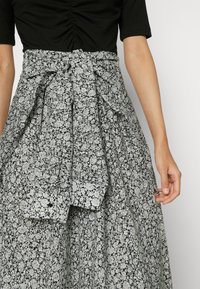 Gros plan d'une personne portant un haut noir froncé et une jupe taille haute noire et blanche à motif floral avec une ceinture en tissu nouée.