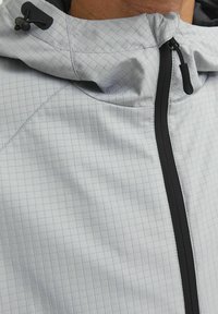 Hellgraue Jacke mit einem Karomuster, Kapuze und einem Frontreißverschluss. Der Stoff hat eine glatte Textur mit dunklen Akzenten am Reißverschluss.