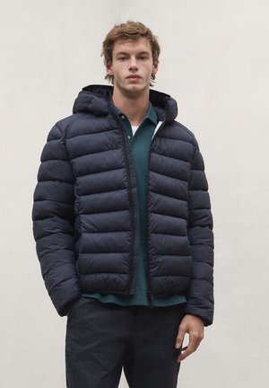Young man standing against a plain wall wearing a black puffer jacket with hood, dark pants, and a green collared shirt.