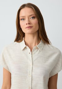 Camicia beige chiara a maniche corte realizzata in lino testurizzato. Presenta una chiusura con bottoni frontale e un colletto classico con sottili linee verticali.