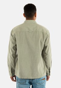 Light green corduroy shirt with long sleeves, buttoned cuffs, and a straight hem. Features a back yoke with a loop detail. Worn with light blue jeans.