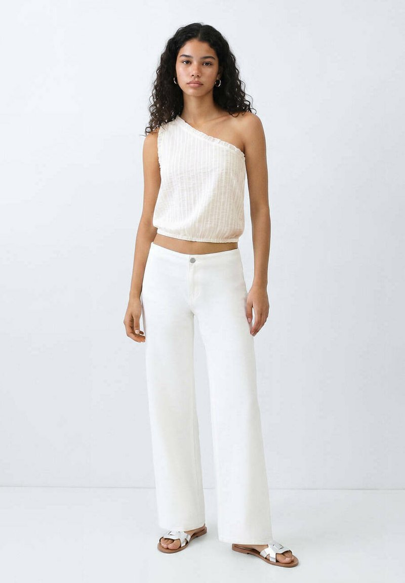 Jeune femme aux cheveux bouclés portant un top blanc à une épaule, un pantalon blanc à jambes larges et des sandales blanches, debout devant un fond uni.