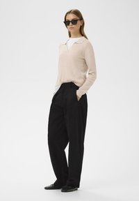 Hellbeiges geripptes Sweater mit V-Ausschnitt, kombiniert mit lockeren schwarzen Hosen. Das Model trägt schwarze eckige Sonnenbrillen und schwarze Schuhe.