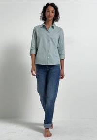Lichtgroene gestreepte blouse met opgerolde mouwen, gecombineerd met blauwe denim jeans. De jeans zijn omgeslagen bij de enkels en worden blote voeten gedragen.
